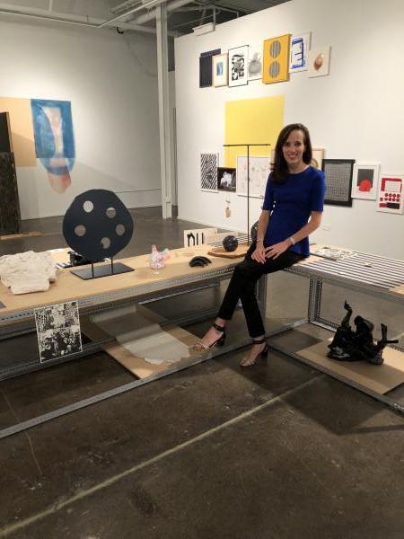 A woman with medium-length brown hair sits on a bench surrounded by various sculptures and works on paper.