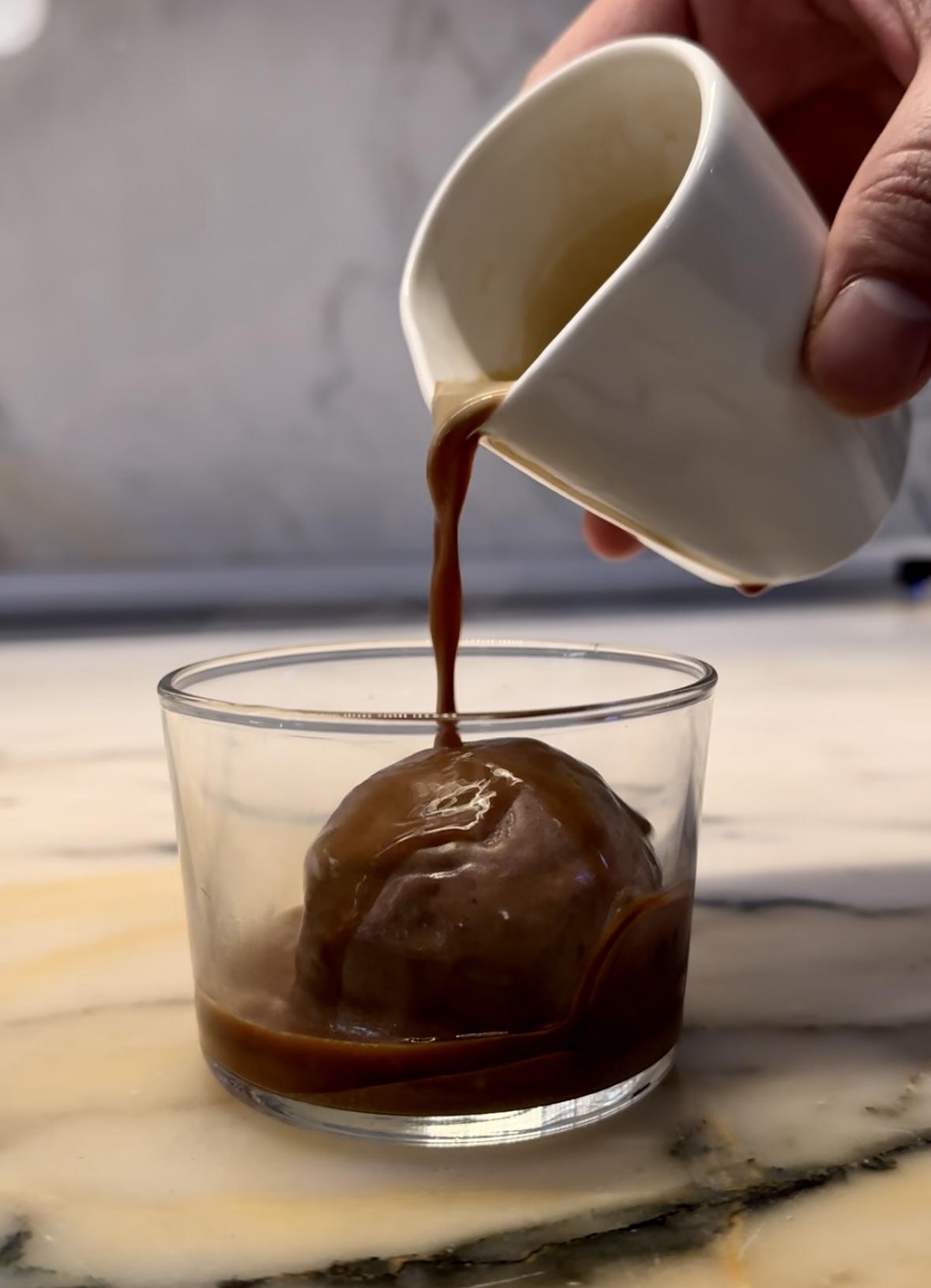 A small carafe of brown liquid is poured onto a scoop of chocolate ice cream within a glass bowl.