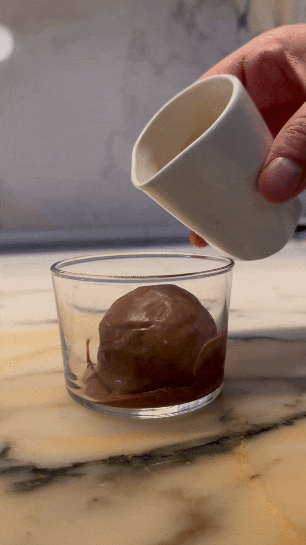 Gif of a small white carafe pouring brown liquid onto a scoop of ice cream. The steam from the mixture fogs up the glass bowl