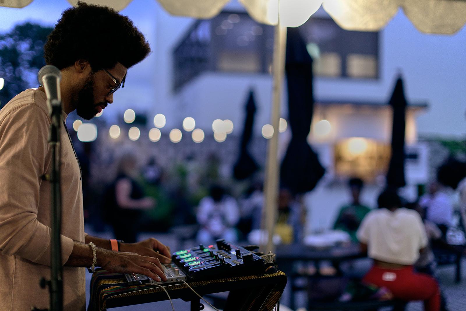Three-quarter view of a man using a soundboard. In the background, groups of people gather at patio tables.