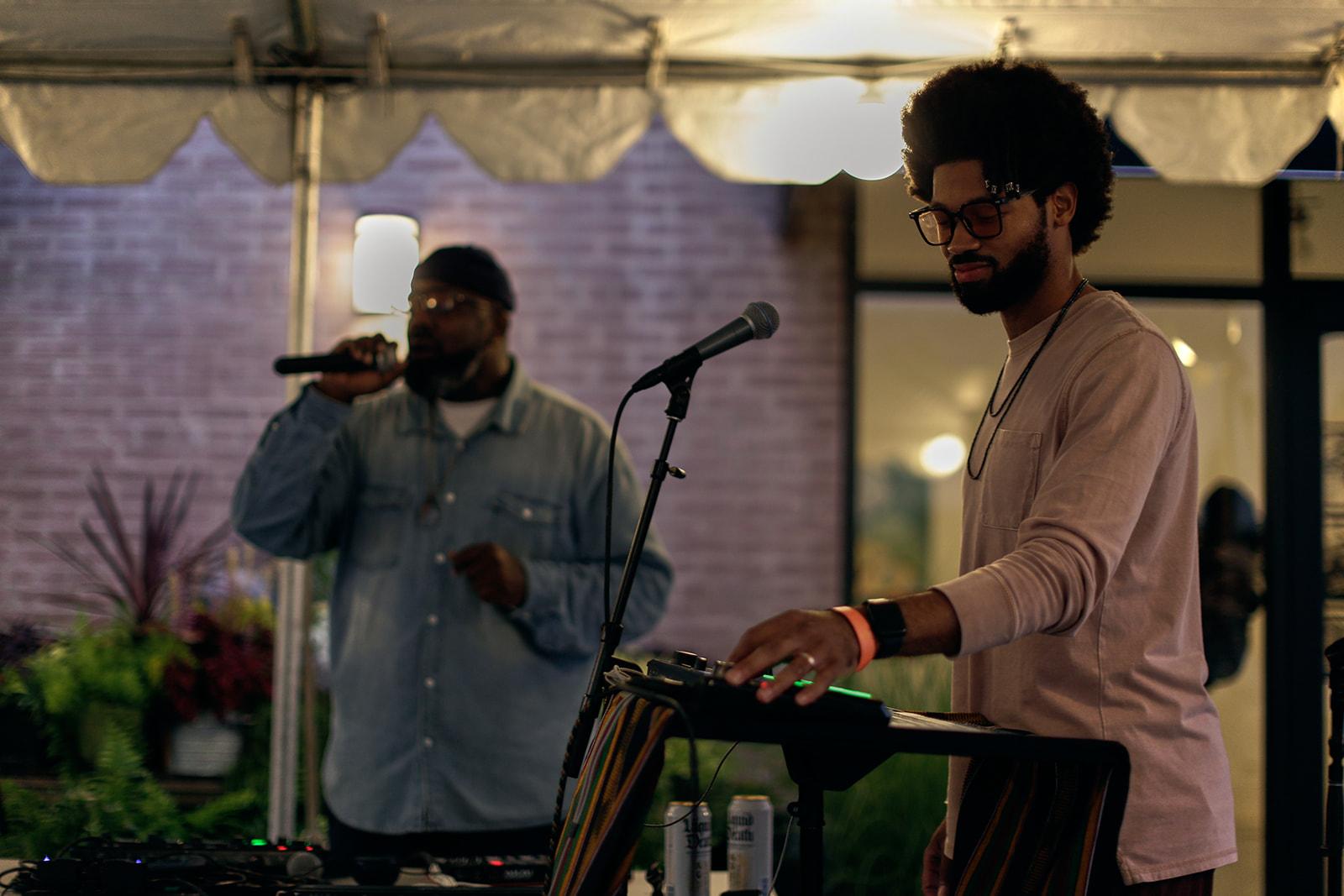 Under a canopy tent a man turns a knob on a soundboard in the foreground. In the background another man holds a microphone up to his mouth.