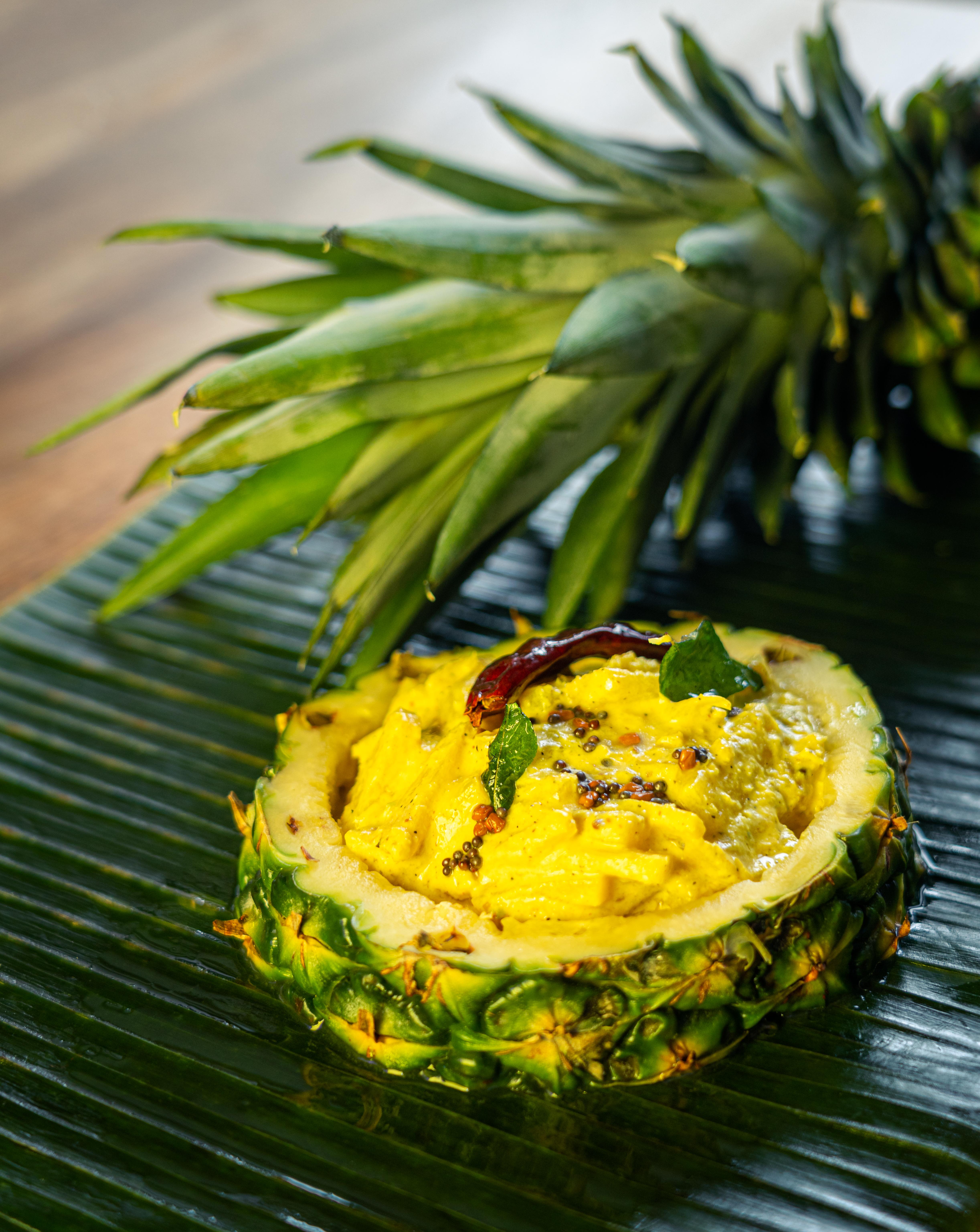 A sectioned pineapple filled with a creamy yellow dip onto of a banana leaf. The crown of the pineapple is placed behind it.