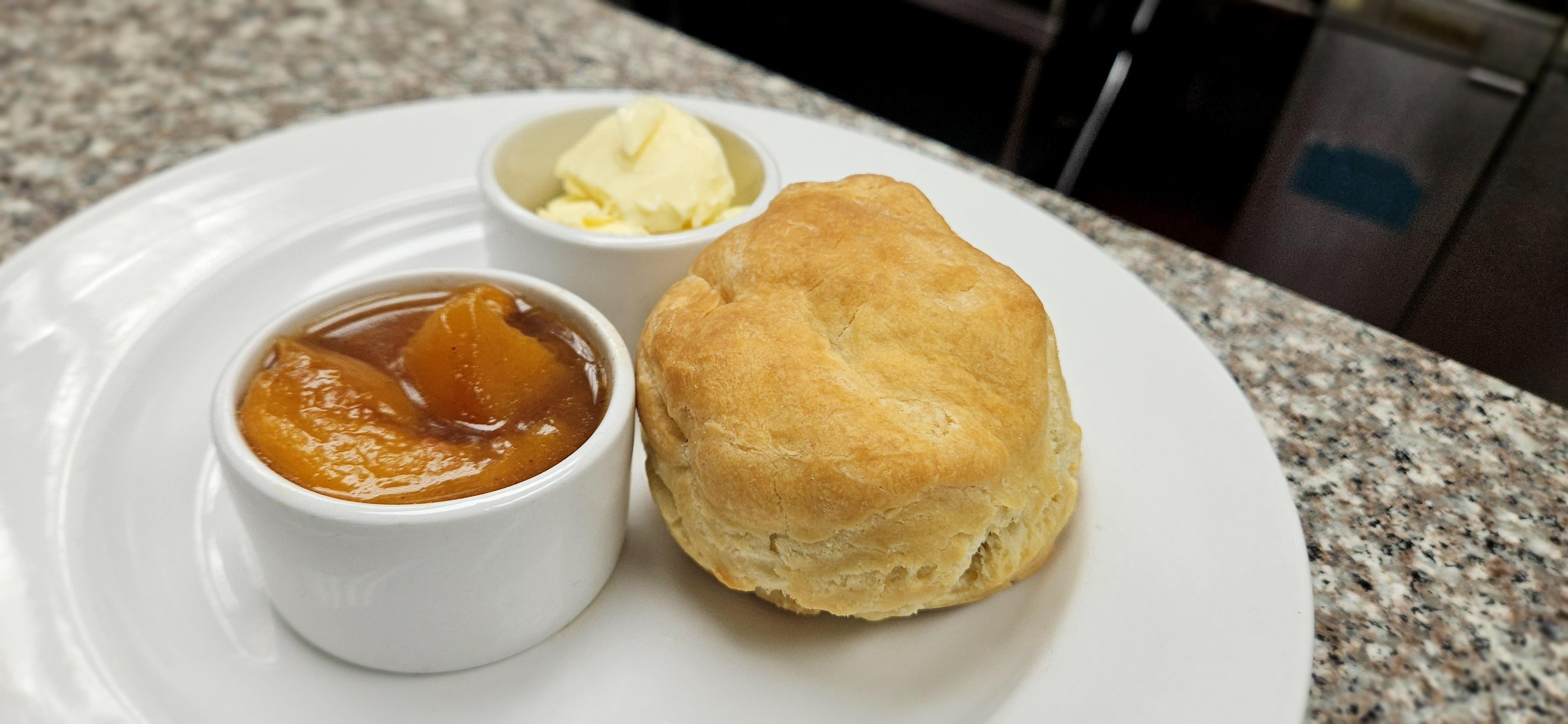 A baked biscuit rests on a large white plate next to two small bowls. One is filled with an orange jelly-like substance, the other is filled with small scoops of butter.