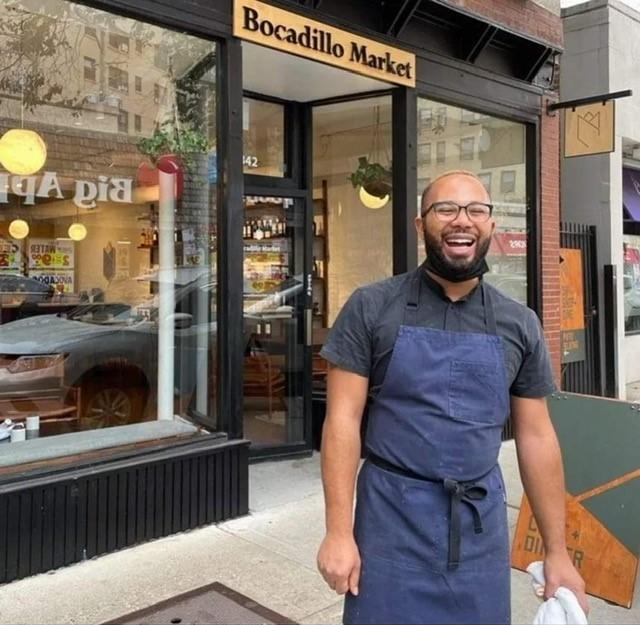 A light-skinned Black man wearing a blue chef's apron is captured mid-laugh outside a building labeled Bocadillo Market.