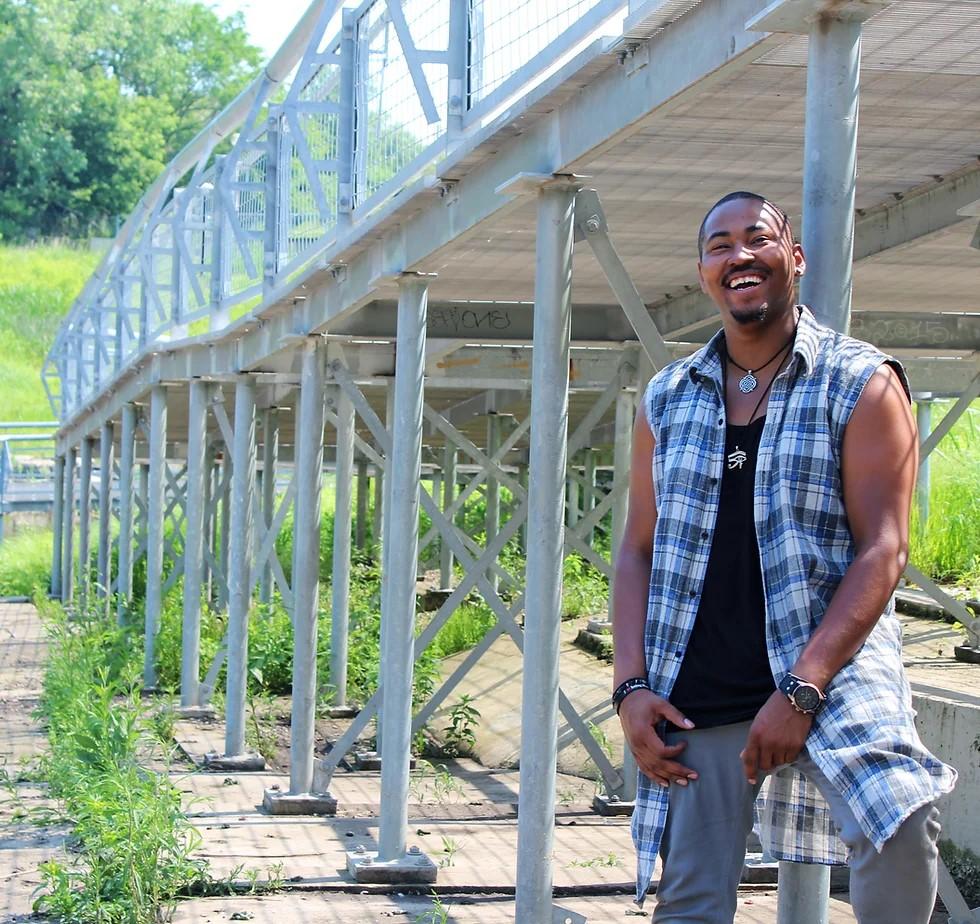 A darker-skinned man in a cutoff button-down flannel smiling widely at the camera poses beside the undergirding of a raised walkway.
