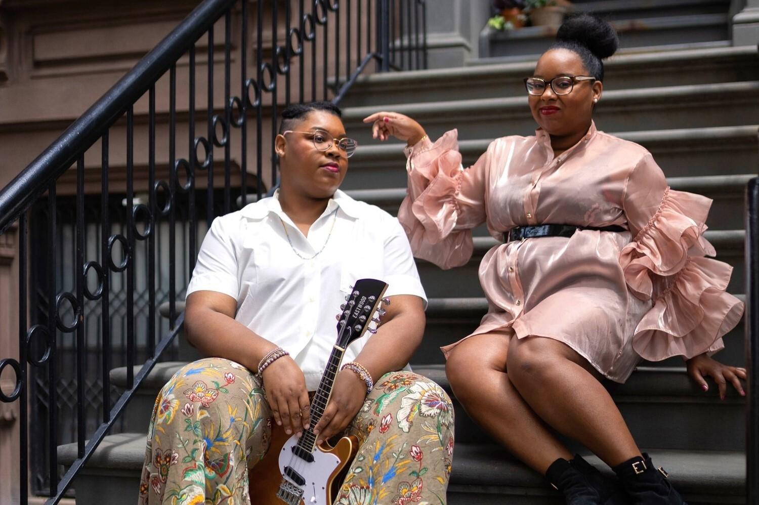 Two Black femmes sitting on the steps of a brownstone building