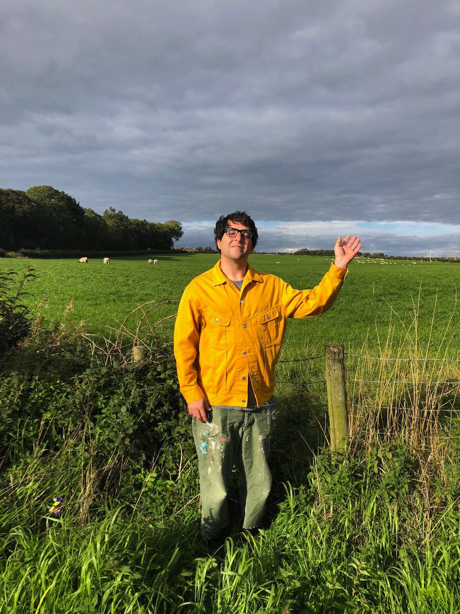 A man waves from the middle of a field near a sheep pasture.