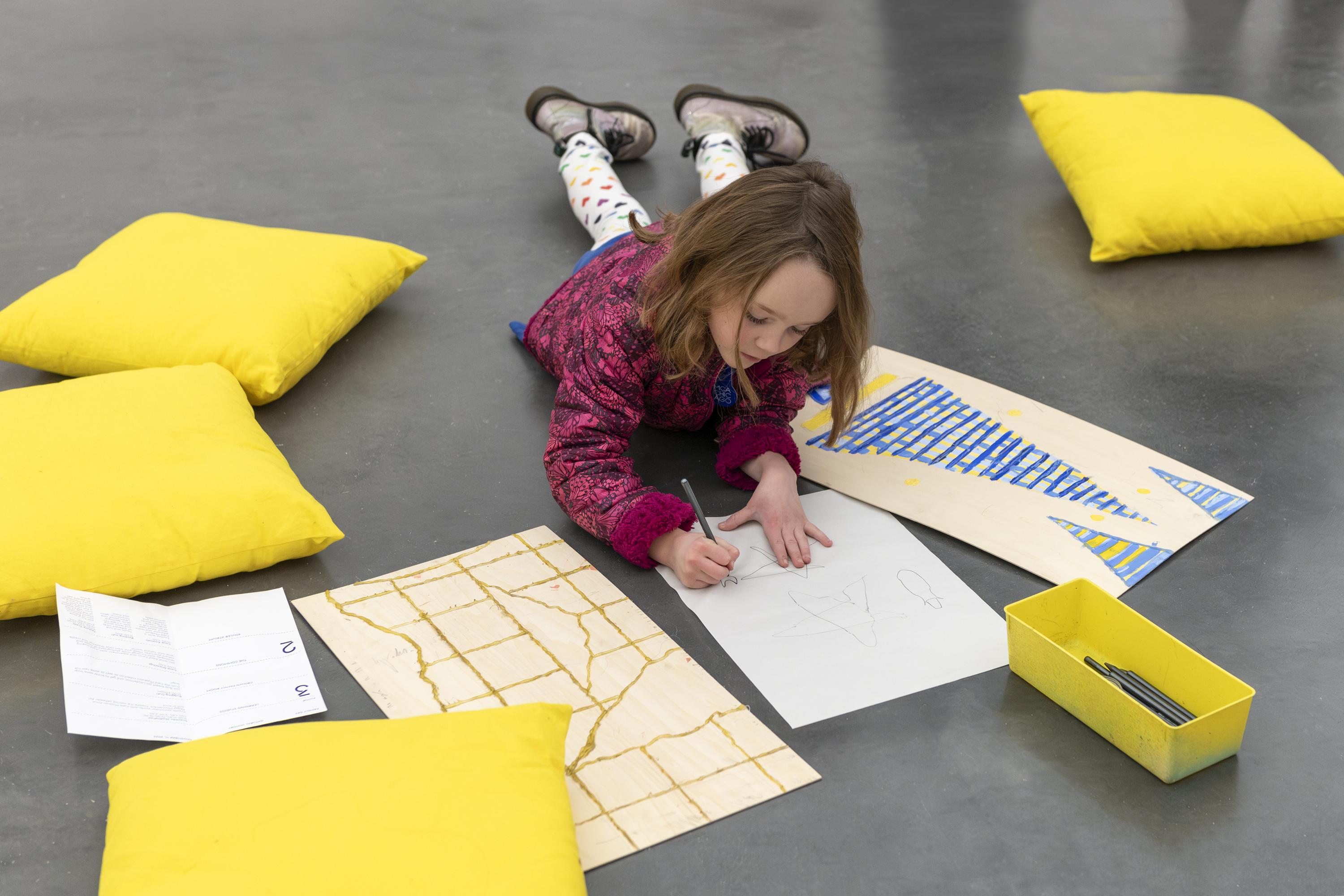 A child lays on a concrete floor while coloring, surrounded by yellow pillows and pieces of paper.