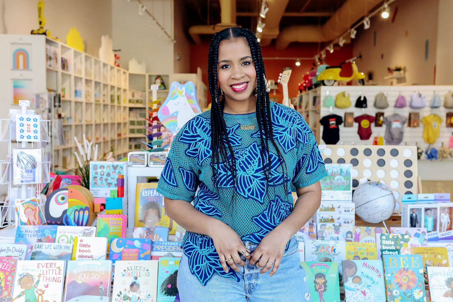 A Black woman poses in front of a display of books in a store, with various clothing and toys in the background.