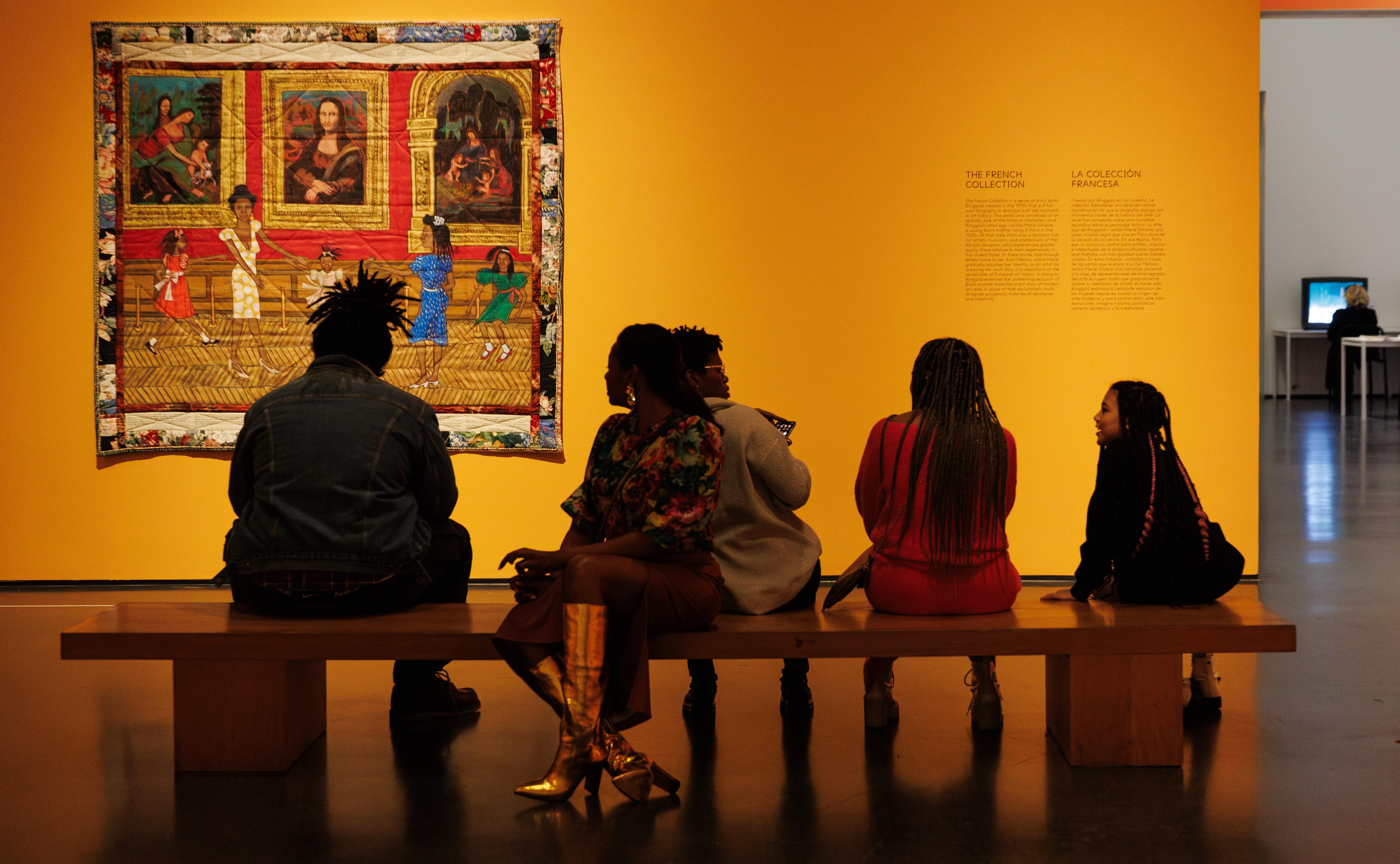 Five people sit on a long wooden bench in an orange gallery space with a quilt hung on the wall.