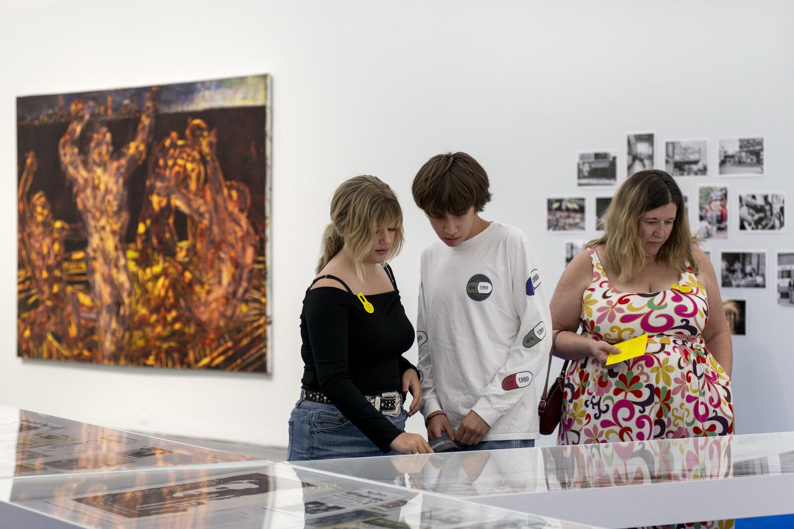 Three people look at a collection of object in display cases. Behind them, several artworks hang on a white wall.
