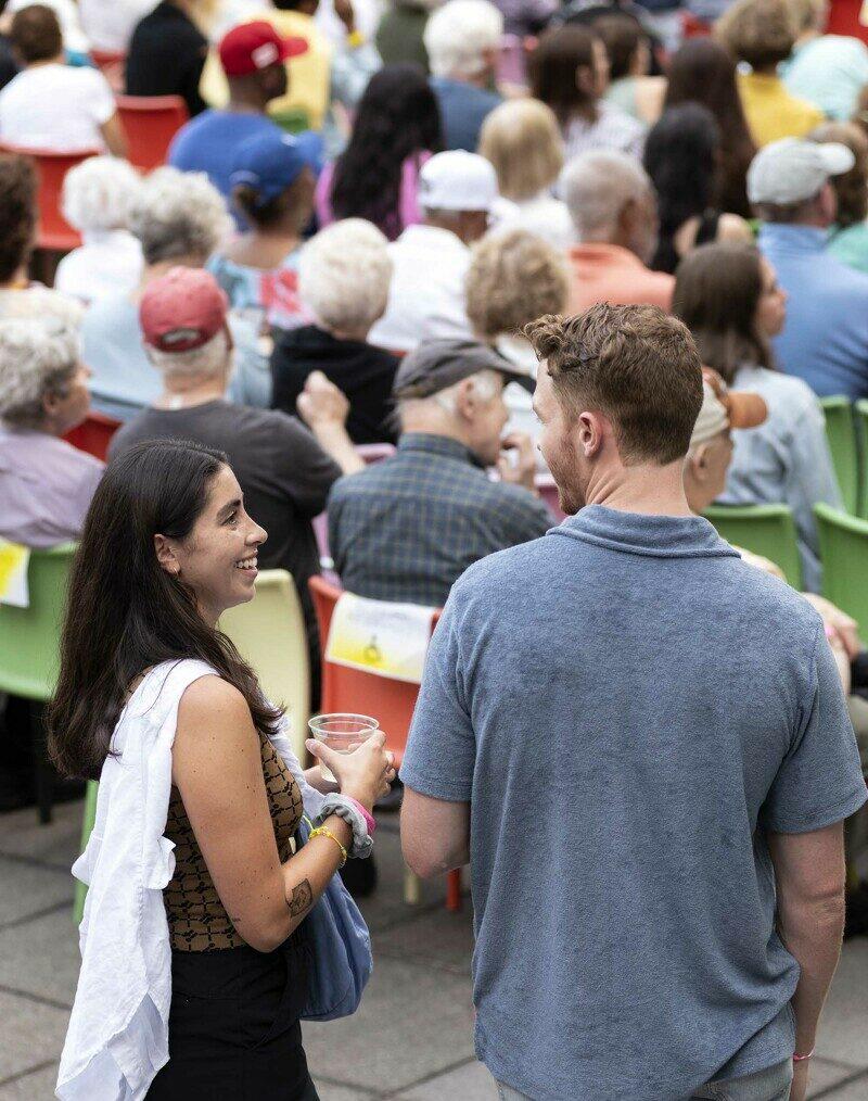 Two people with their backs to the camera interact in front of a large crowd of seated people.