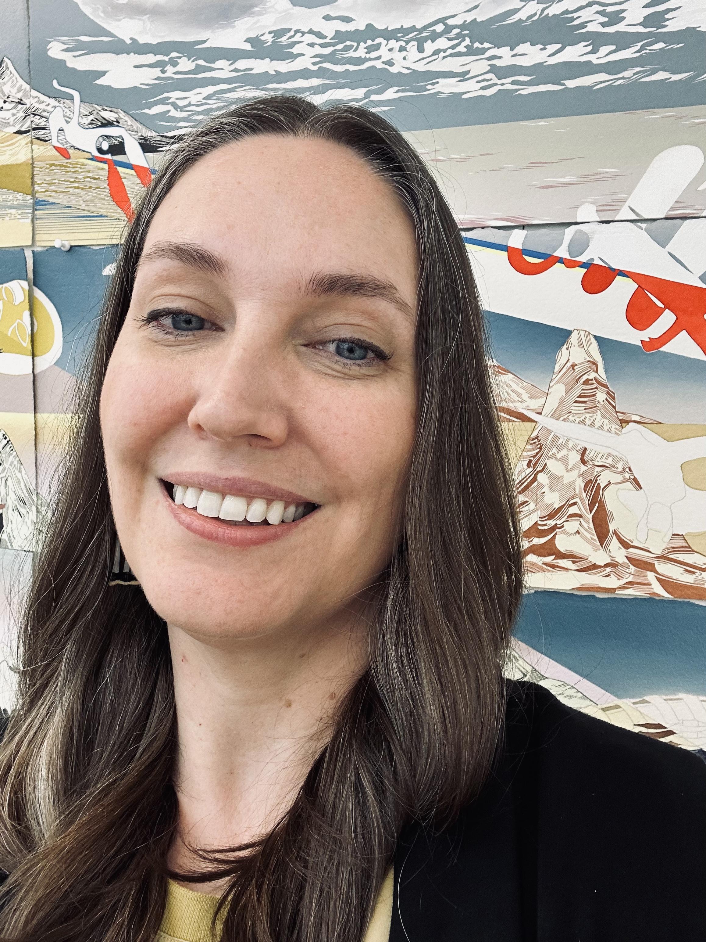 Selfie-like portrait of a woman with long brown hair posing in front of a large artwork.