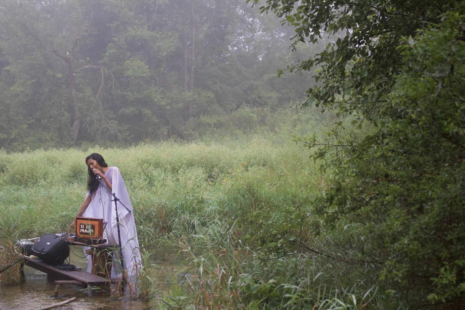 A woman in a white dress performs into a microphone amid wetlands