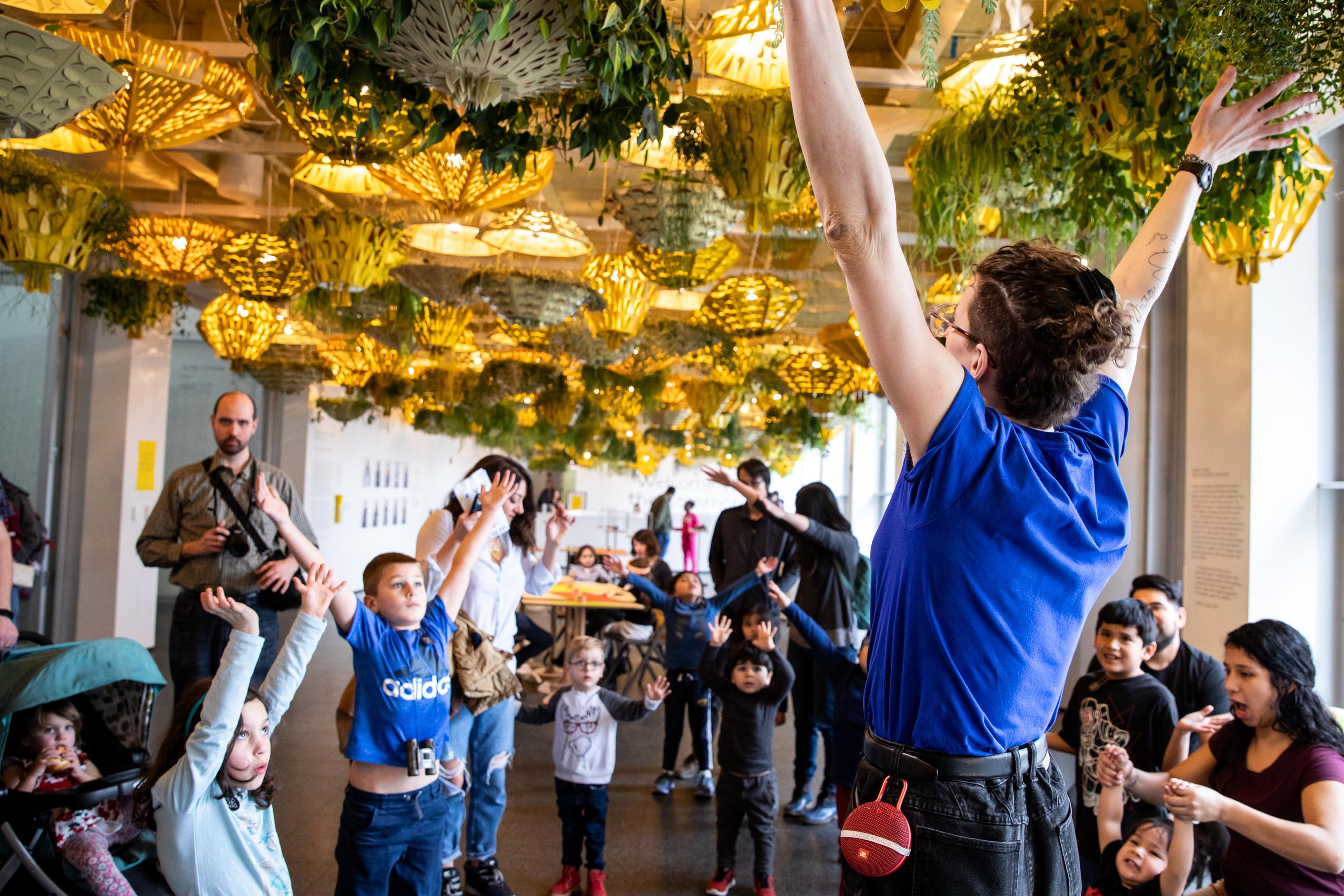 A group of kids raise their arms, mirroring the pose of an adult standing in front of them in a room full of planters hanging from the ceiling