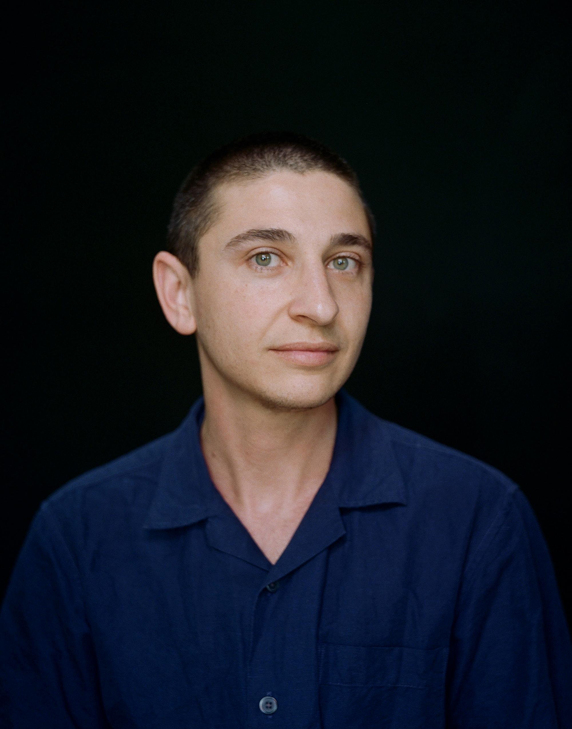 Portrait of a person with closely cut hair and a blue button down looks directly into the camera with their striking green eyes