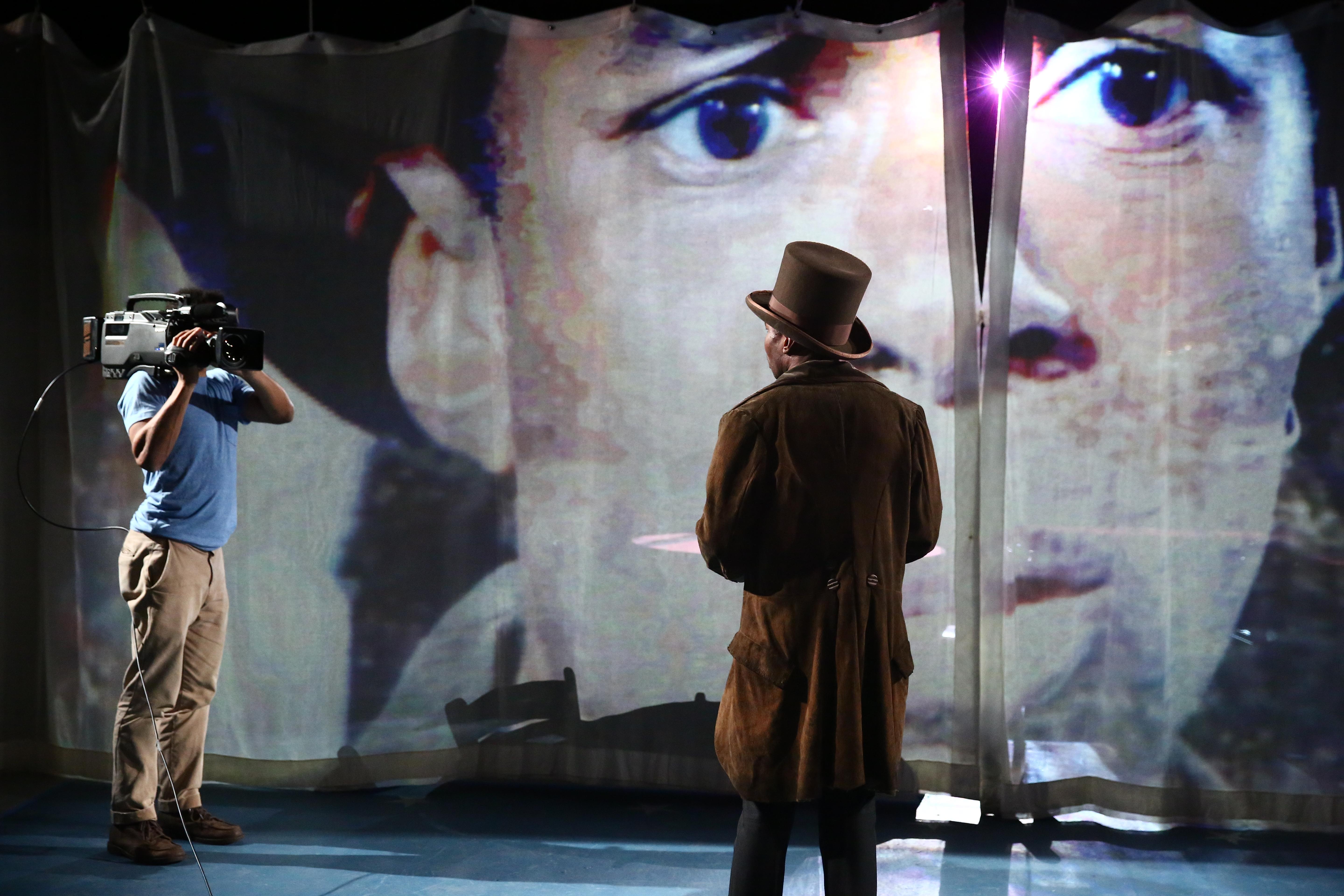 An actor in a top hat faces a person with a video camera in front of a large projection of a concerned looking man