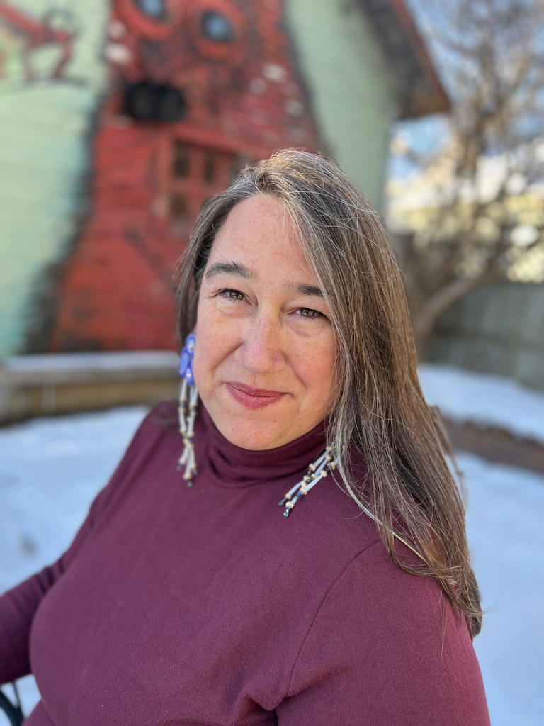 A woman with long grey-brown hair poses outside in a purple turtleneck