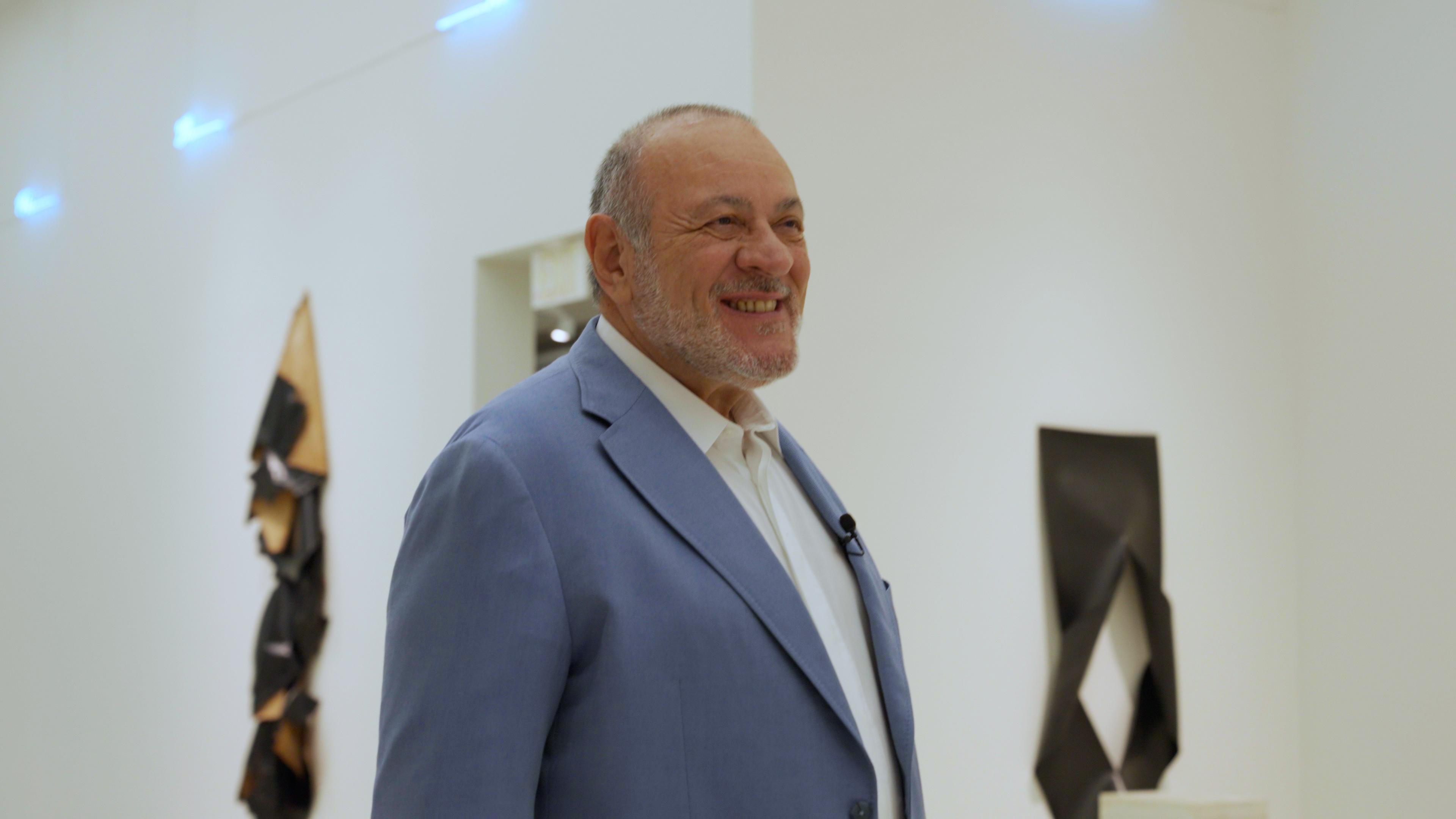 A light-skinned man in a grey suit smiles in a white gallery space with two artworks on the wall behind him