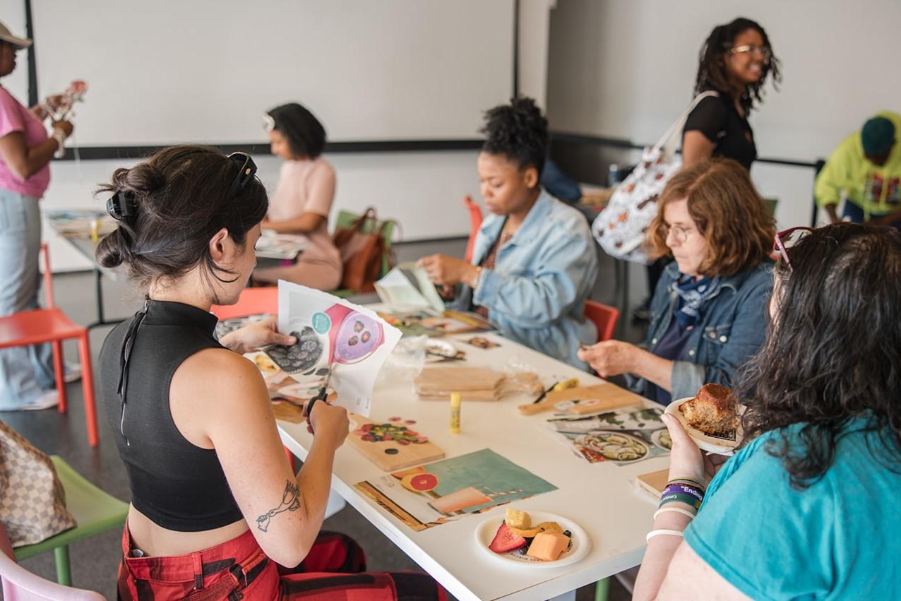 A group of people sit at a table with magazine pages scattered across it. Some are working on cutting out images from these pages.