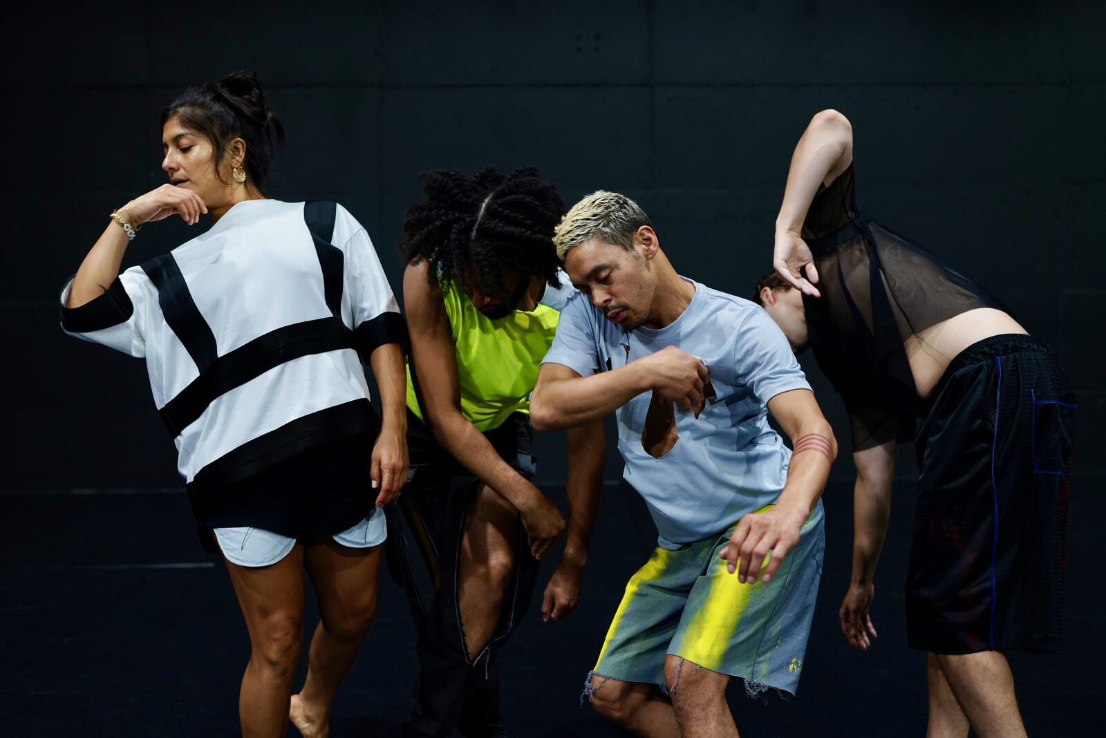 A group of people of various ethnicities holding various positions against a black brick wall.