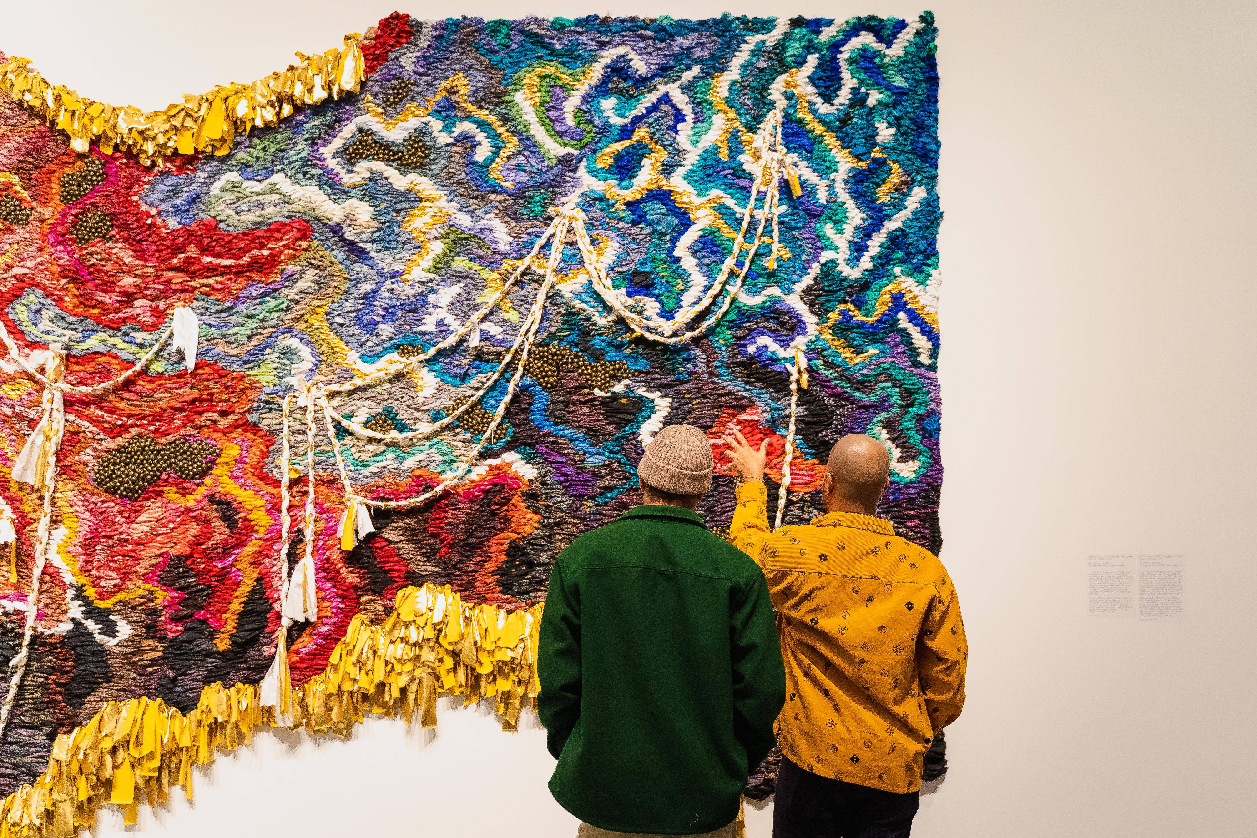 Two people look at a colorful weaving hanging on a wall