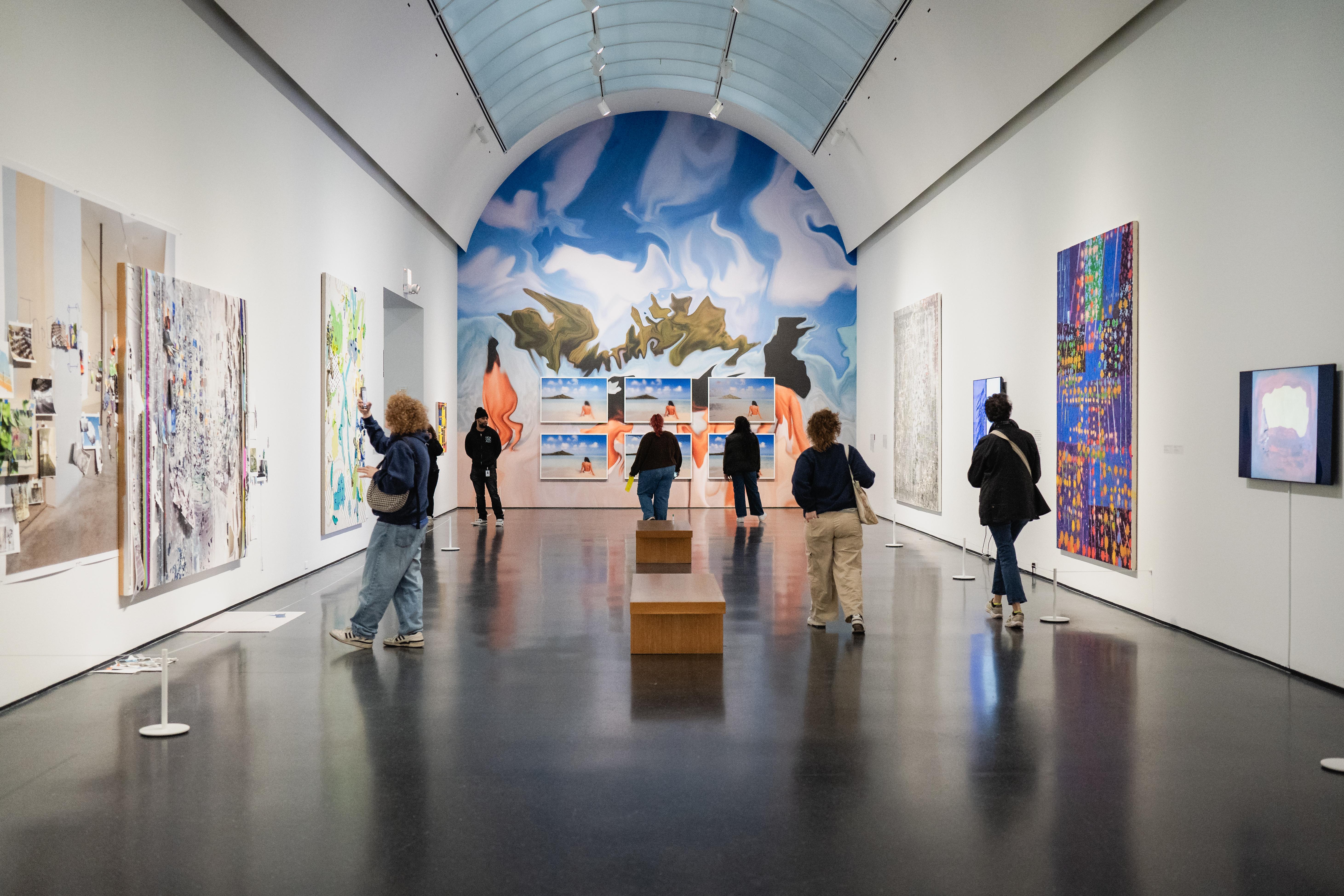 A scattering of people in an airy white gallery space with a cloudlike mural on the back wall and artworks hanging on the two facing walls