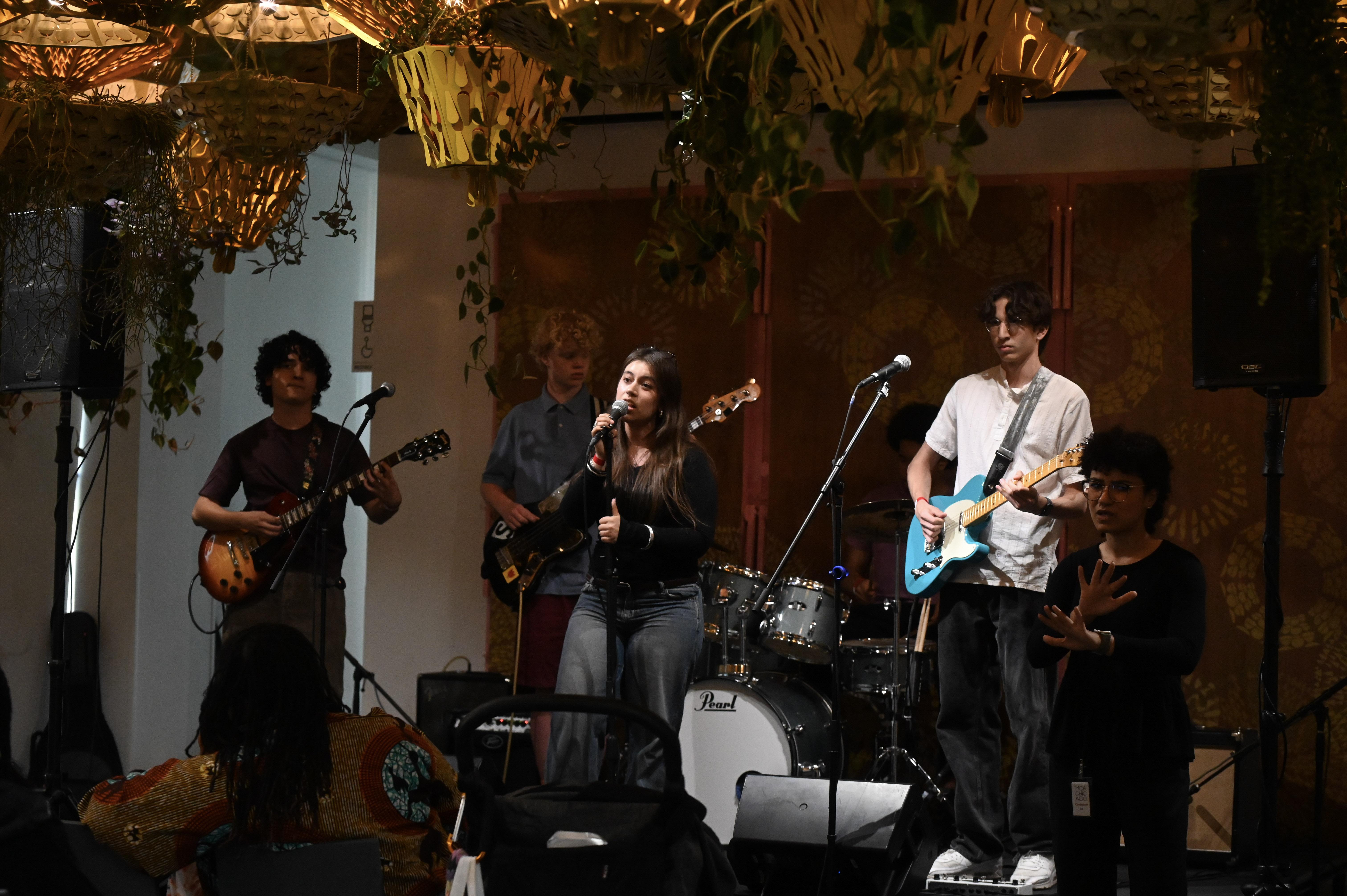 A five piece band plays on a small stage in a room filled with hanging plants. An ASL interpreter stands just to the right of them.