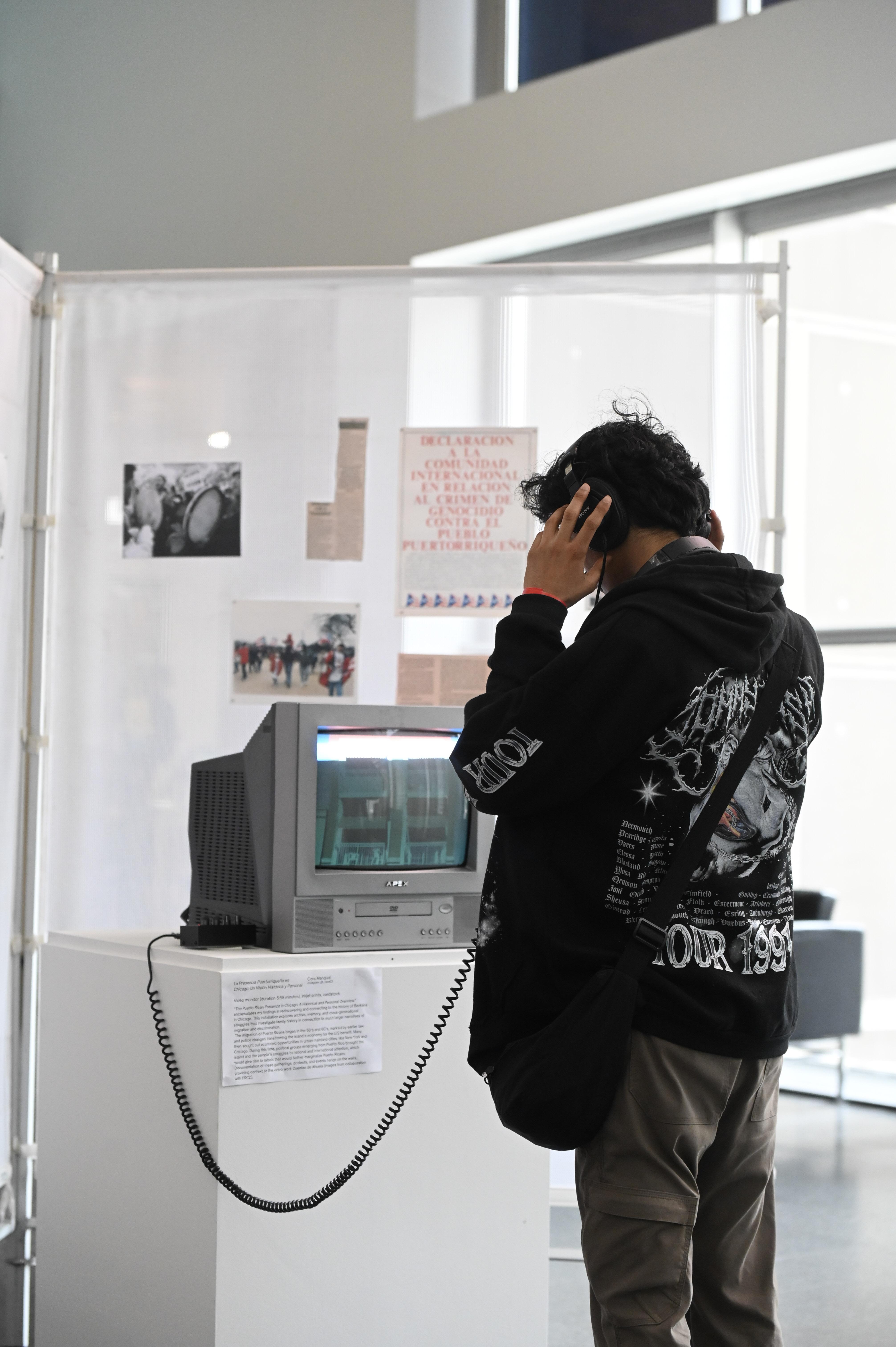 A person in a black hoodie holds analog headphones to their ears. The headphones are attached to an old tv with a built-in DVD player.