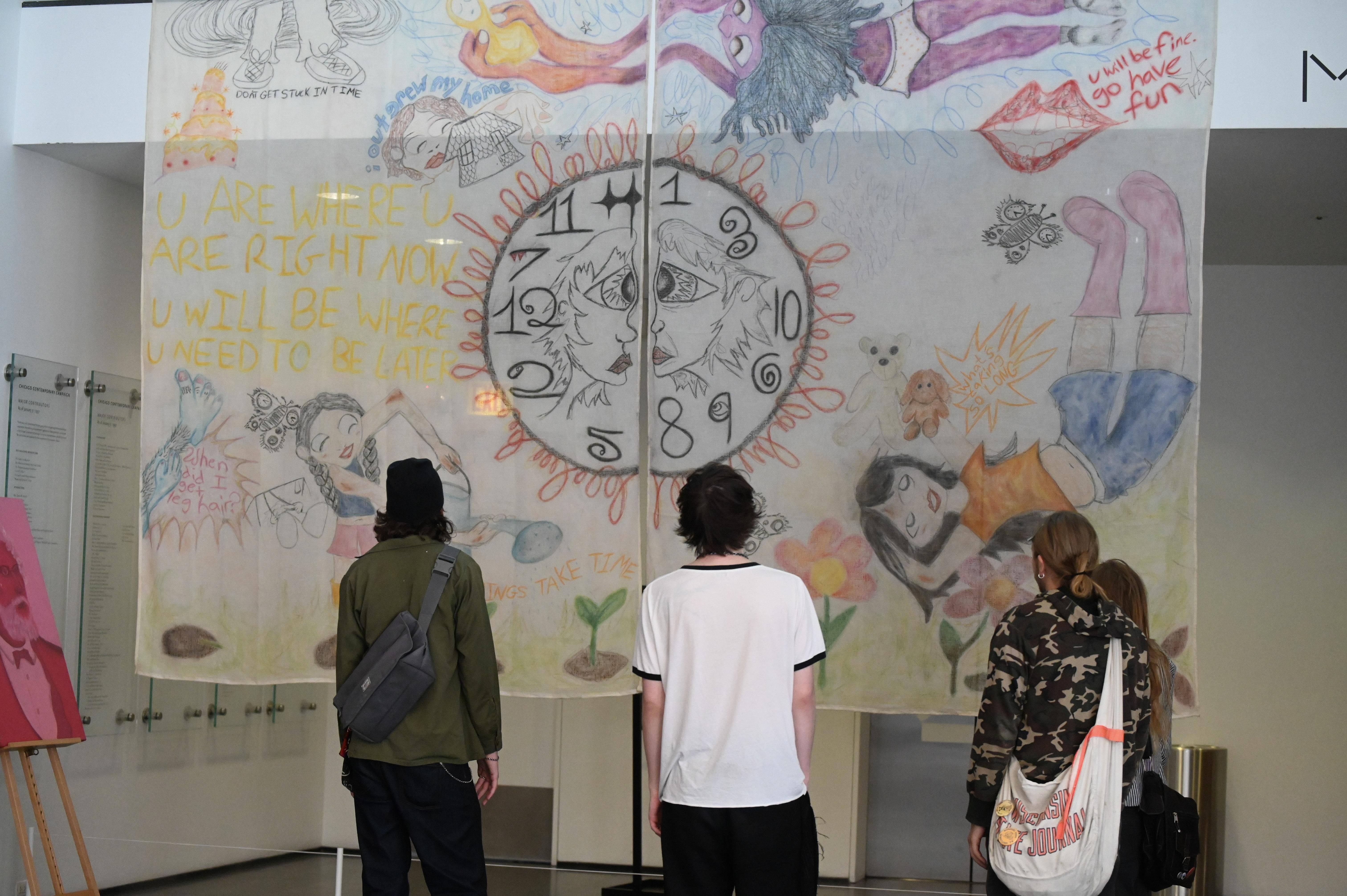 Three people look at two sheets hanging from a balcony with drawings of figures floating around a clock with two faces on it