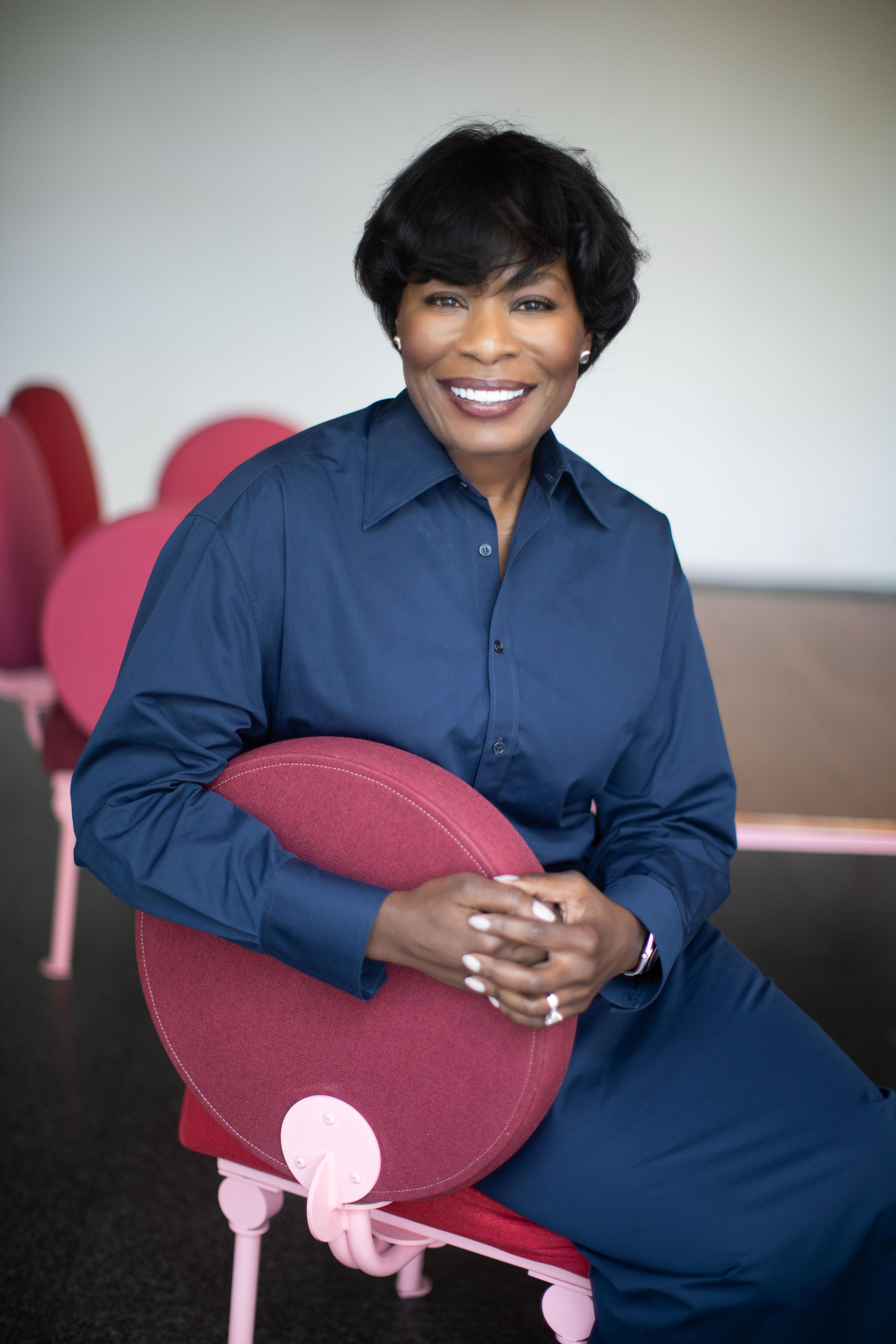A woman with medium-dark skin and short dark hair smiles at the camera while sitting in a red chair, her hands clasped around the back of it