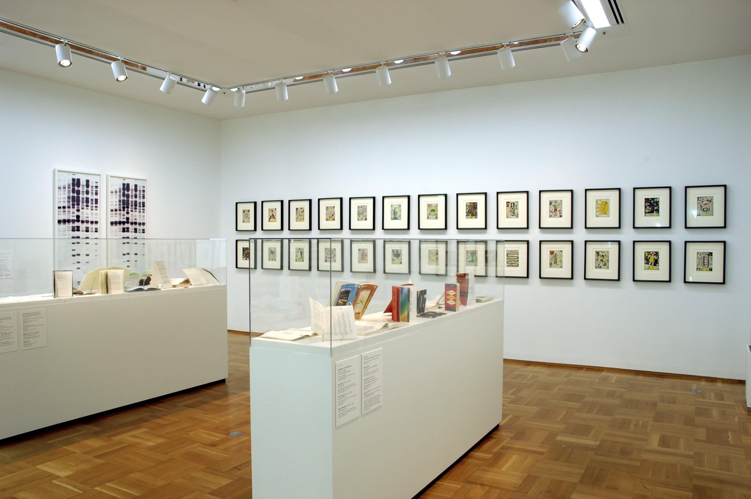 Two glass vitrines hold several books each in a room with artworks on two adjoining walls