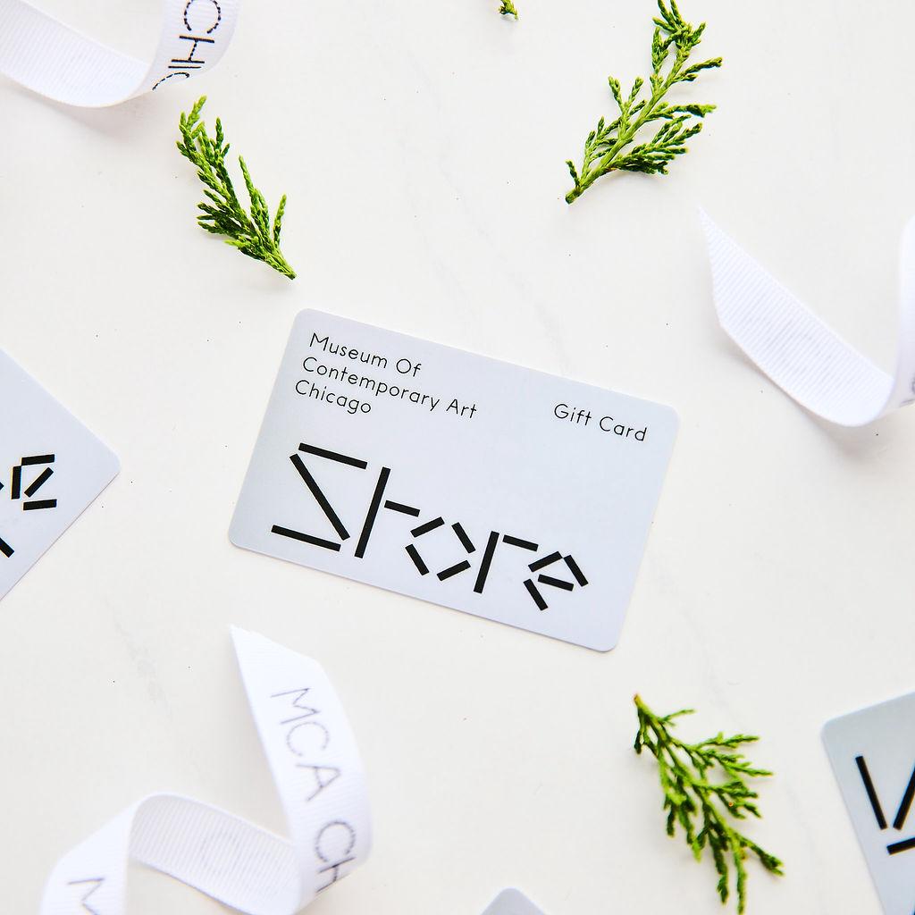 A white gift card labeled Museum of Contemporary Art Chicago Store on a white background with springs of evergreen and white MCA Chicago ribbon surrounding it