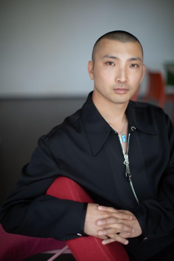 Portrait of a male-presenting person with very close-cut hair dressed in a black zipup polo shirt posed with their arm resting on the back of a red chair